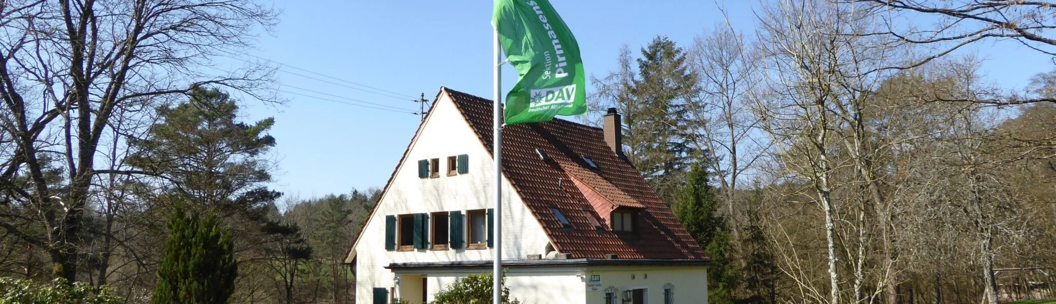 Das Bild zeigt die Frontansicht des Rudolf-Keller-Hauses. Die Fahne der Sektion weht am Fahnenmast vor dem Haus. | © DAV