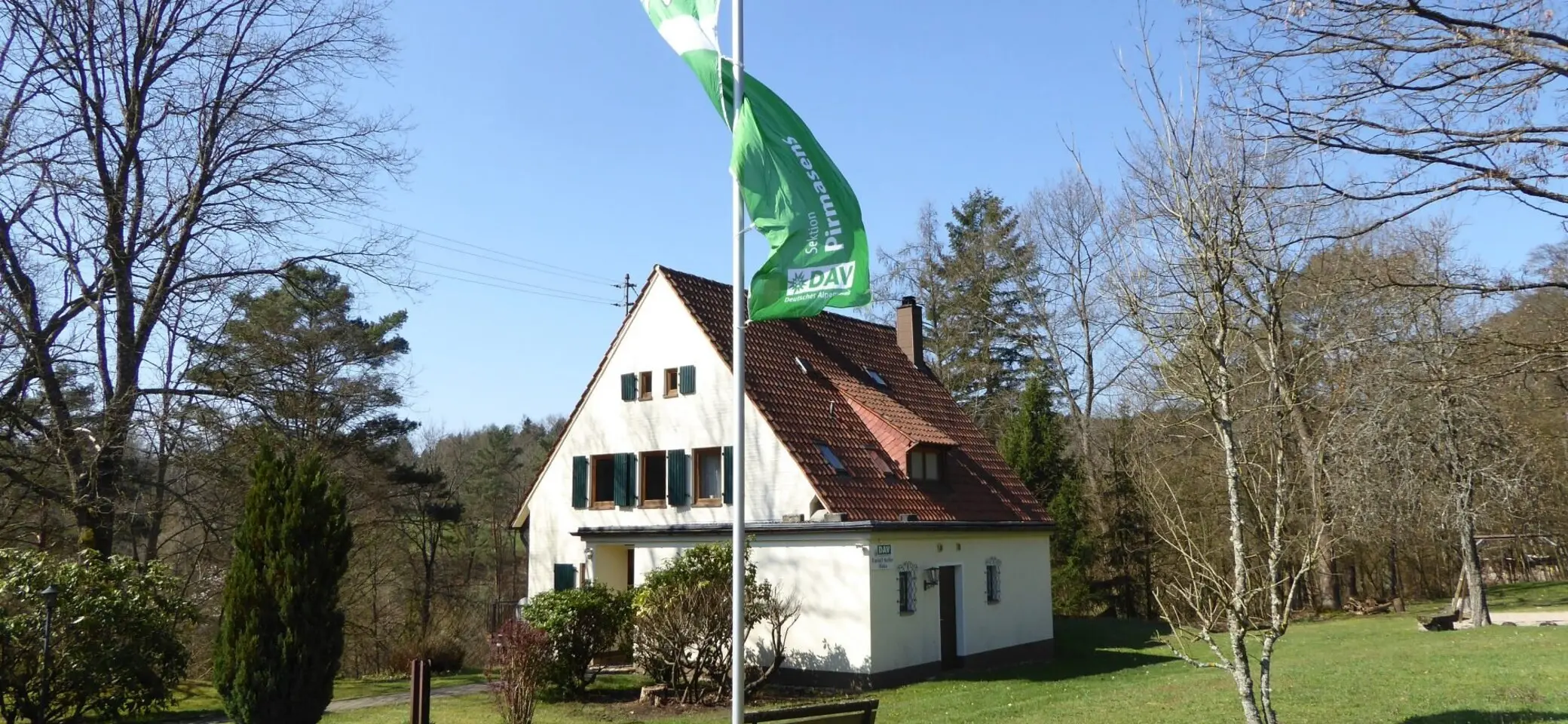 Das Bild zeigt die Frontansicht des Rudolf-Keller-Hauses. Die Fahne der Sektion weht am Fahnenmast vor dem Haus. | © DAV