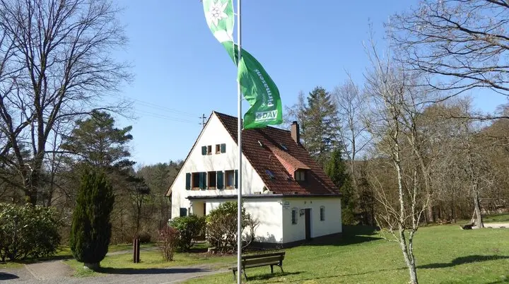 Das Bild zeigt die Frontansicht des Rudolf-Keller-Hauses. Die Fahne der Sektion weht am Fahnenmast vor dem Haus. | © DAV