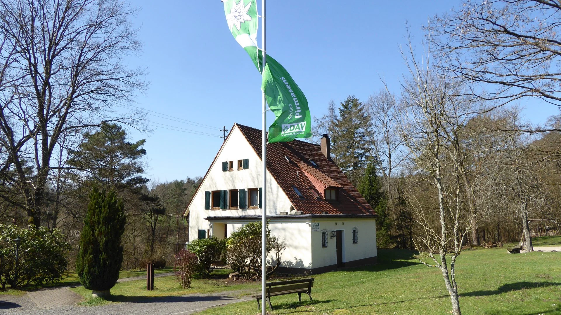 Das Bild zeigt die Frontansicht des Rudolf-Keller-Hauses. Die Fahne der Sektion weht am Fahnenmast vor dem Haus. | © DAV