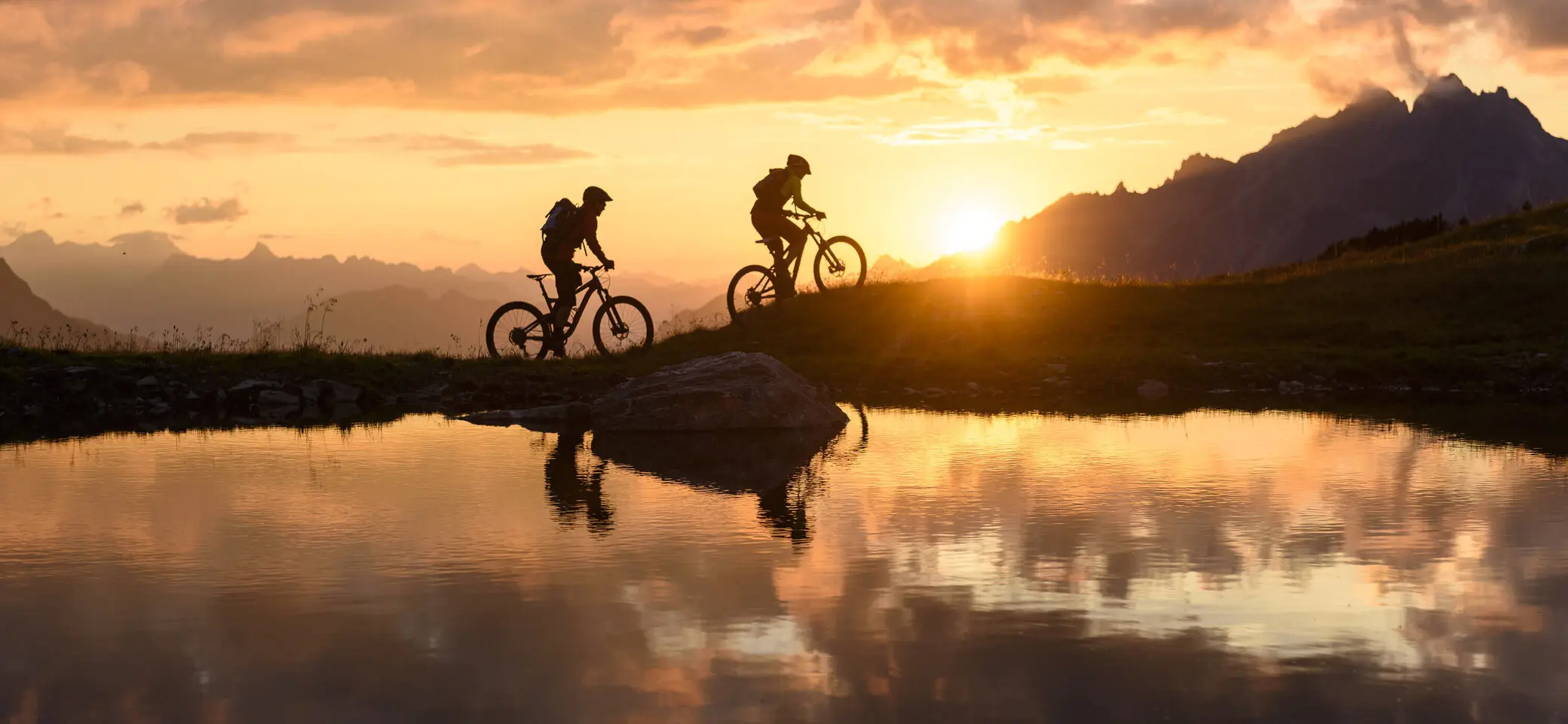 Zwei Personen fahren im Sonnenuntergang mit ihren Mountainbikes vor einer Bergkulisse. | © DAV/Wolfgang Ehn